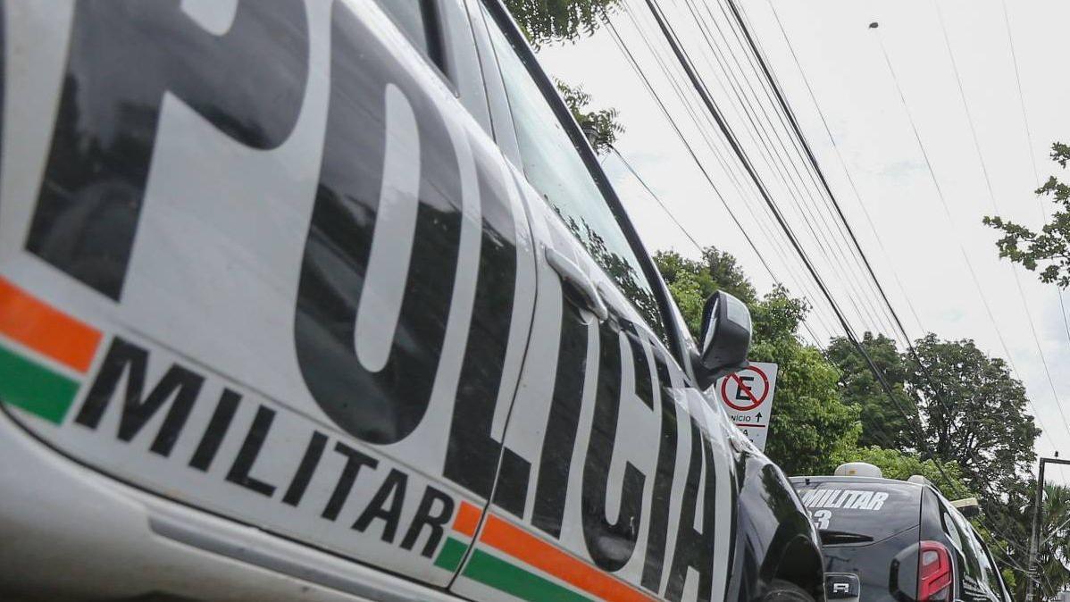 A imagem mostra duas viaturas da Polícia Militar do Ceará, brancas e adesivadas nas cores preto, laranja e verde, em um local com árvores e uma placa de Proibido Estacionar.