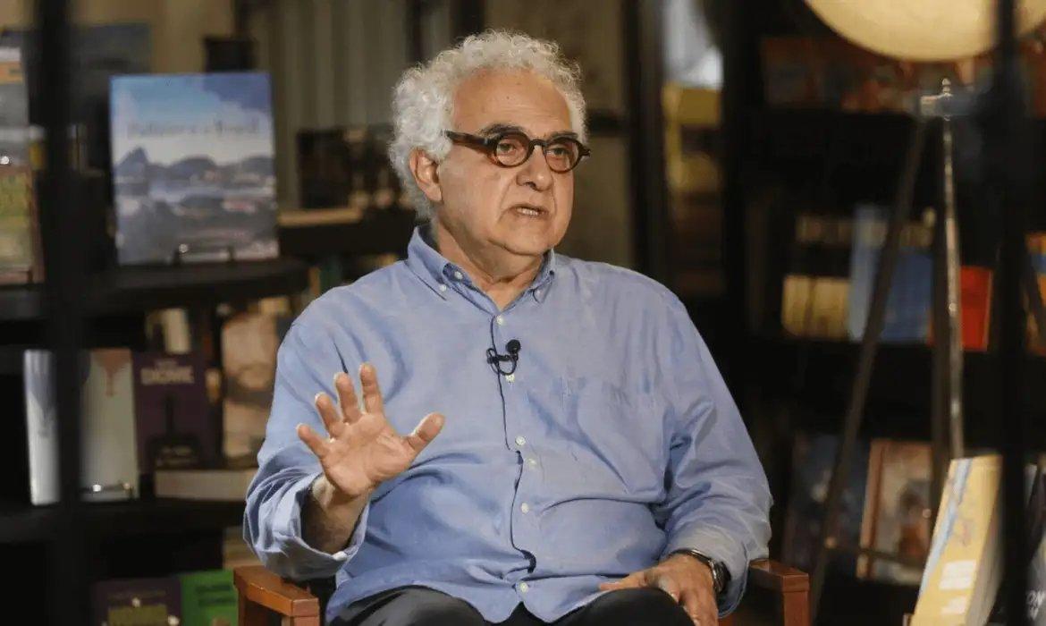 Milton Hatoum, homem pardo de cabelos brancos, sentado em uma cadeira de escritorio. Cenário de fundo mostra livros.