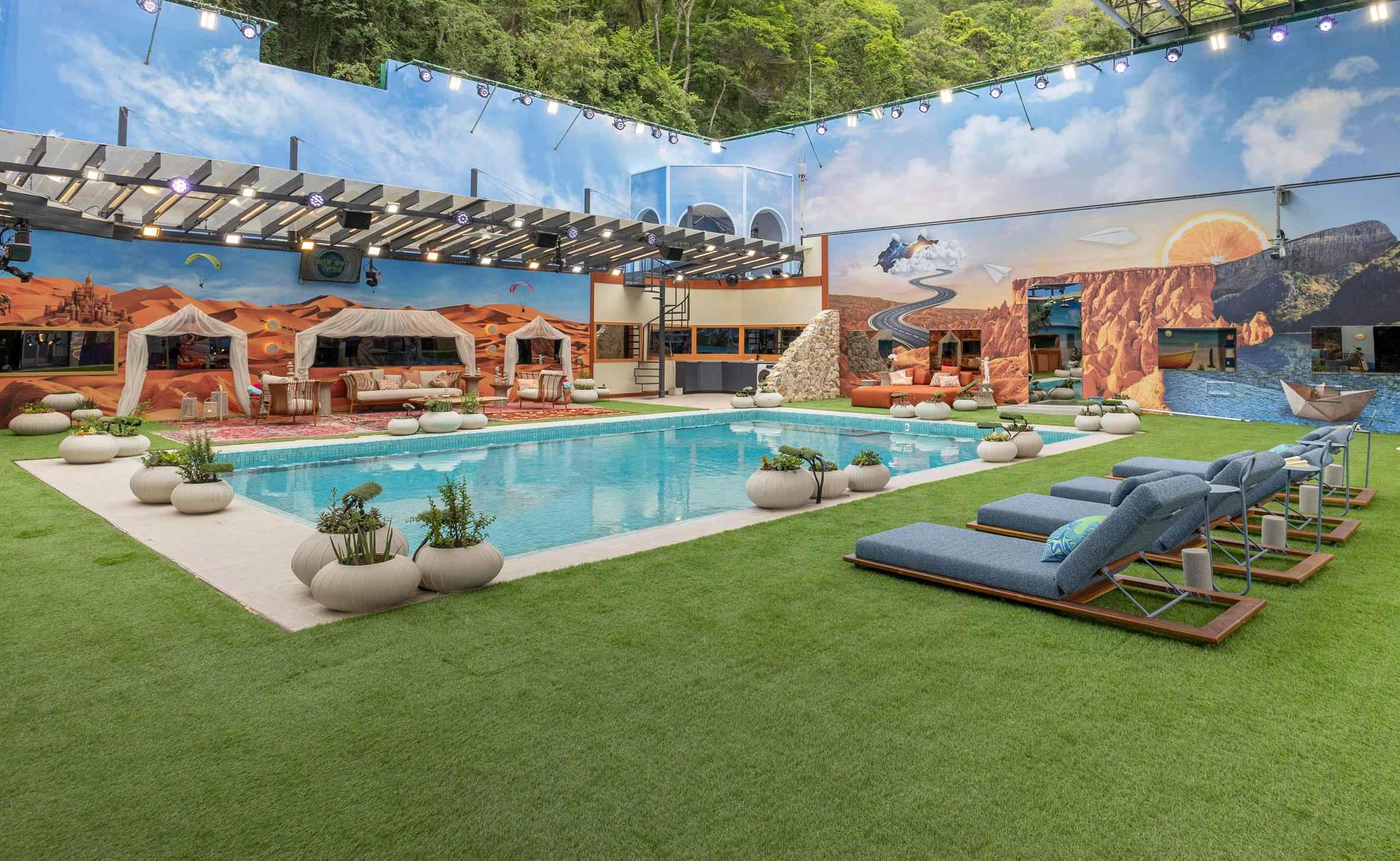 Ampla área externa de lazer com piscina, gramado artificial, espreguiçadeiras e grandes murais temáticos de deserto com tendas e montanhas com estrada sinuosa.