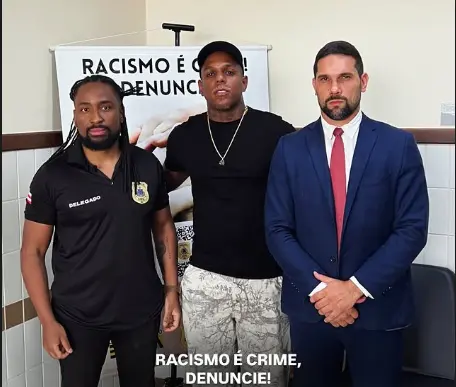 Três homens posam lado a lado em frente a um banner educativo sobre o combate ao racismo. À esquerda, um homem negro com dreadlocks veste uma camisa polo preta com a palavra Delegado; ao centro, um homem negro de boné e camiseta preta; e à direita, um homem branco de terno azul e gravata vermelha. O banner ao fundo e uma legenda na parte inferior reforçam a mensagem: Racismo é crime, denuncie!.