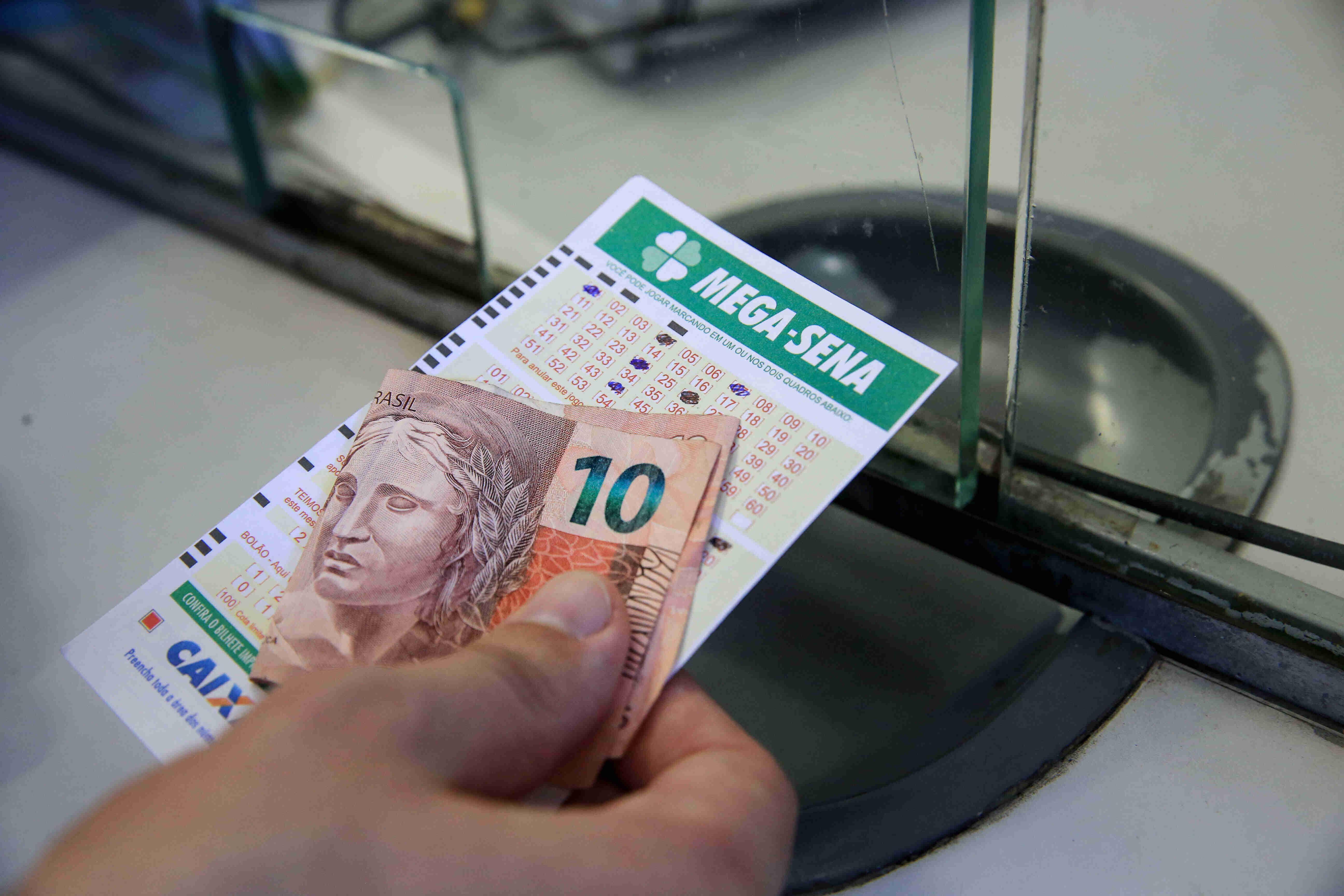 Mão com cartela da Mega Sena e uma nota de R$ 10 reais em mãos.