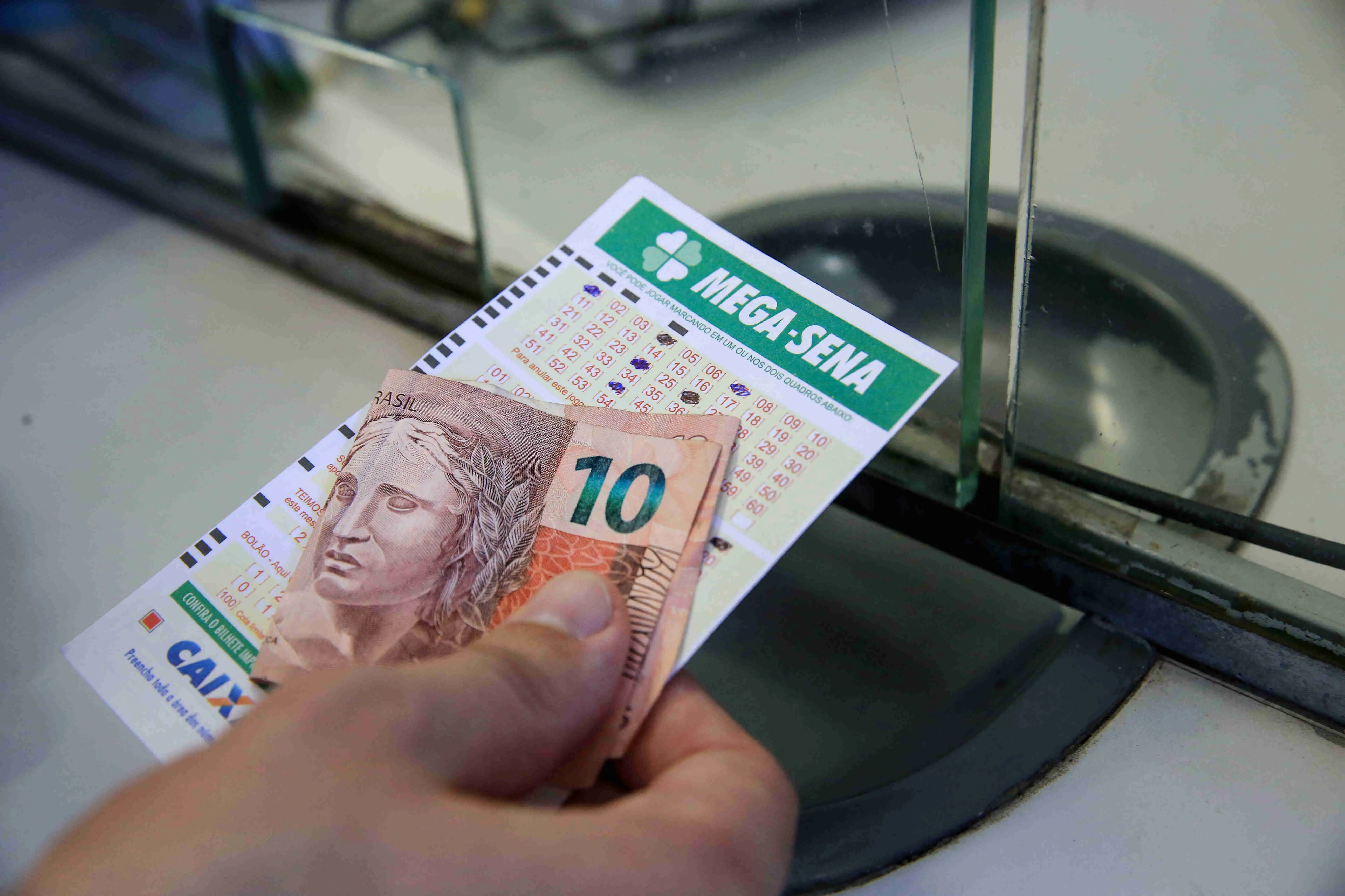 Mão com cartela da Mega Sena e uma nota de R$ 10 reais em mãos.