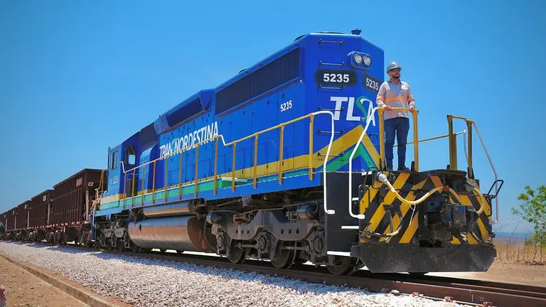 Locomotiva azul e verde da Transnordestina puxa vagões de carga em ferrovia no Nordeste; trabalhador com EPI está sobre a frente do trem durante operação em área aberta e ensolarada.