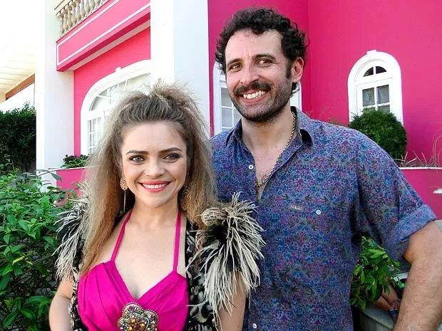 Homem e mulher posam lado a lado em área externa, em frente a um casarão histórico de fachada rosa com janelas brancas; ambos vestem roupas coloridas e estão cercados por vegetação em ambiente urbano.