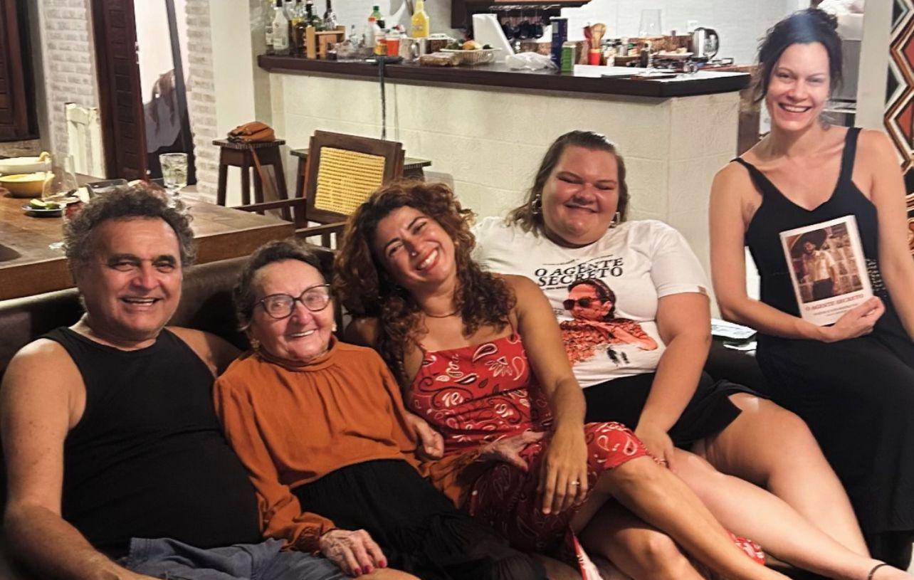 Parte do elenco de 'O Agente Secreto', incluindo a atriz Tânia Maria, sorri sentado em um sofá. O grupo está reunido em um ambiente interno acolhedor com balcão de cozinha ao fundo, transmitindo uma atmosfera de celebração e proximidade.