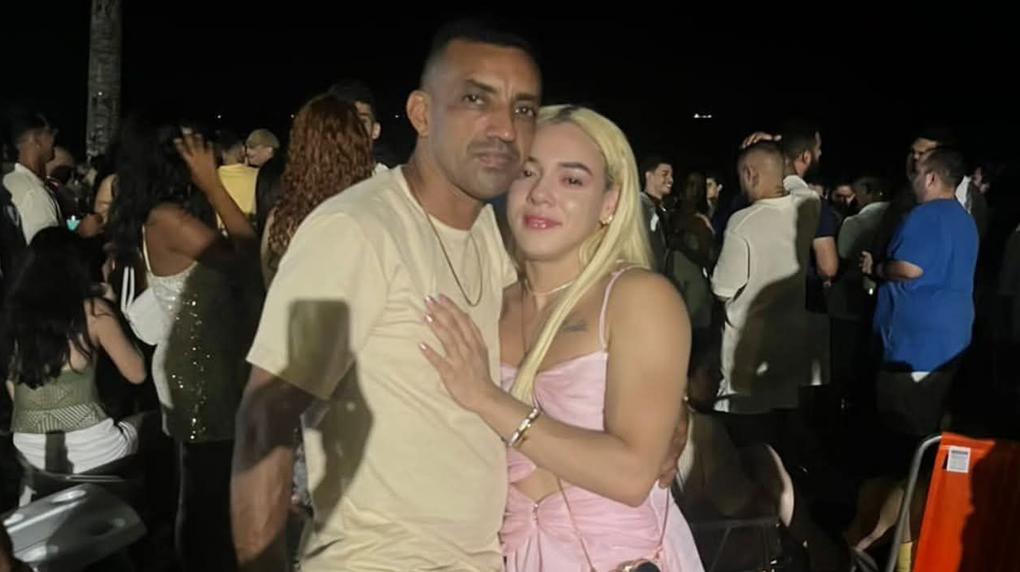 Foto de homem e mulher abraçados em festa com diversas pessoas ao redor.