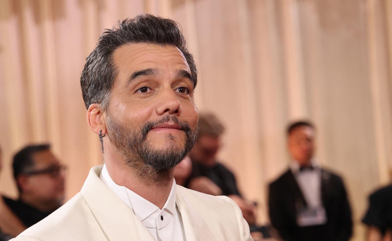 O ator Wagner Moura aparece em close-up, vestindo um traje formal branco impecável e um brinco de argola escura na orelha direita. Ele exibe uma expressão serena e um leve sorriso, com o olhar direcionado para cima contra um fundo desfocado de tons quentes e figuras em trajes de gala.