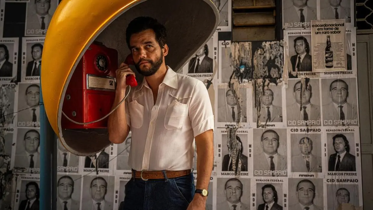 Wagner Moura ao lado de um orelhão vermelho vestindo camisa branca de manga curta.