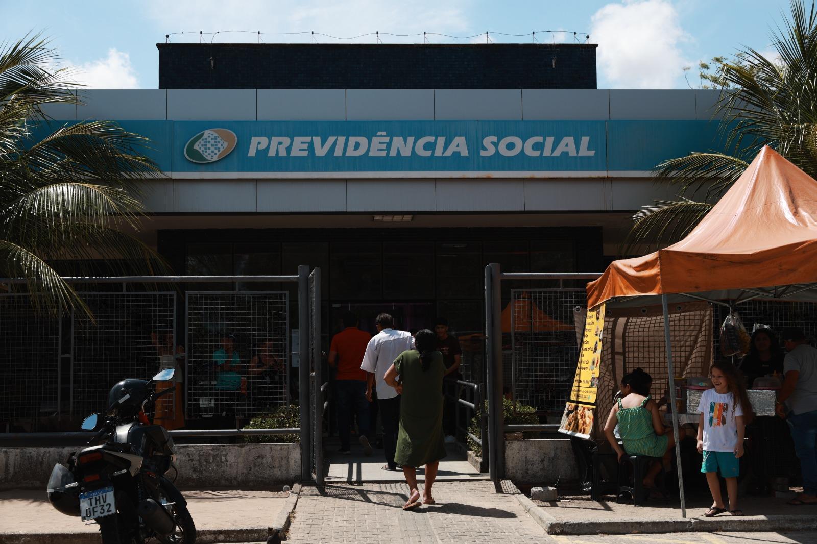 Fachada de uma agência da Previdência Social sob luz solar forte, com uma placa azul de identificação no topo. Na entrada, pessoas caminham em direção à porta protegida por grades, enquanto à direita há uma barraca de lanches com clientes e uma criança sorridente.