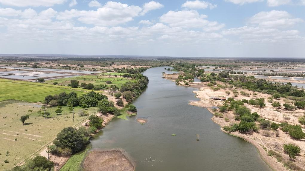 Vista aérea do Rio Jaguaribe, no Ceará, serpenteando por uma paisagem com campos verdes, áreas de cultivo e tanques de aquicultura em suas margens.