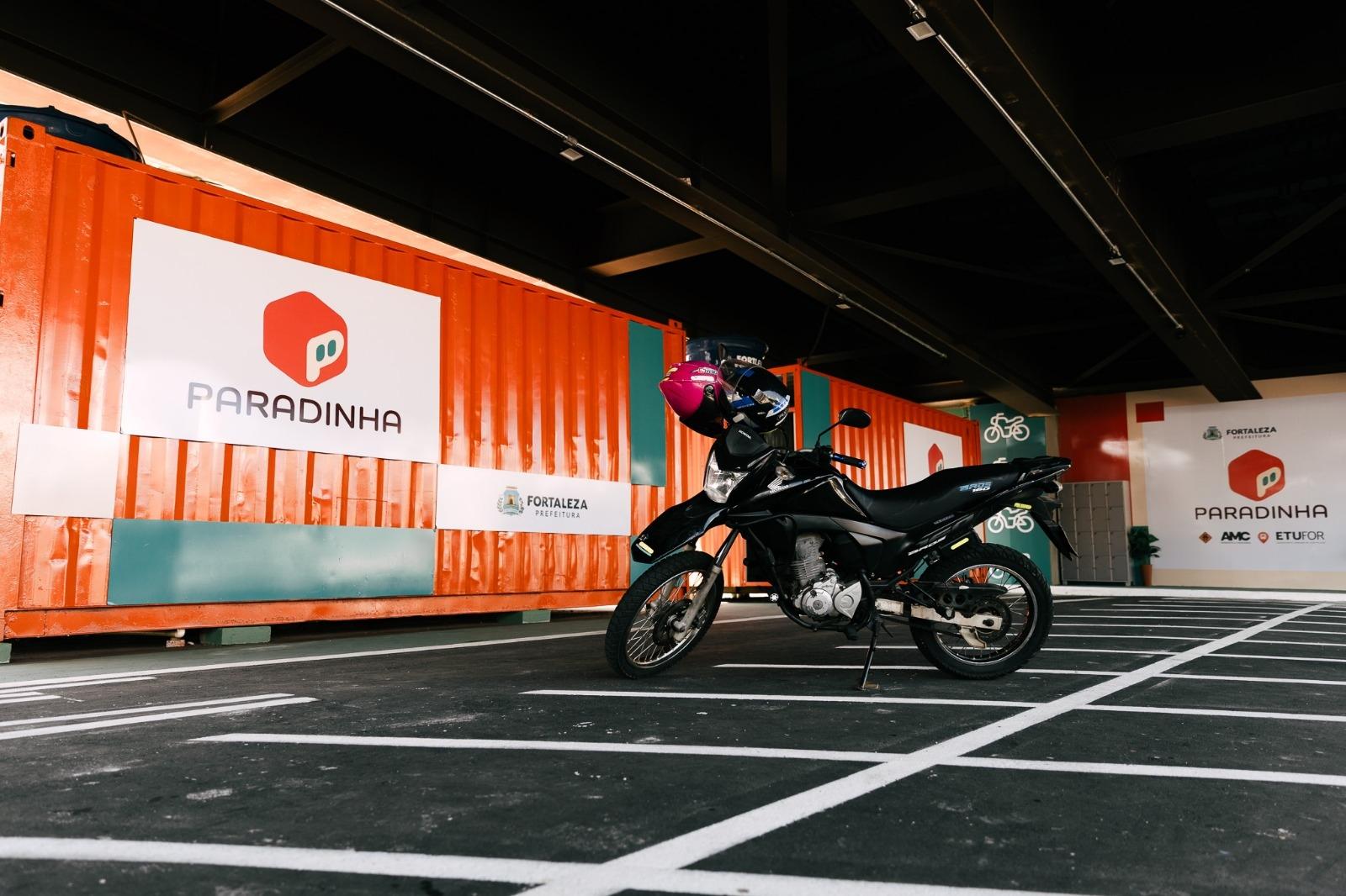 Motocicleta estacionada no projeto Paradinha, em Fortaleza. Contêineres laranjas ao fundo exibem marcas da Prefeitura, AMC e ETUFOR, sinalizando o ponto de apoio para motociclistas.