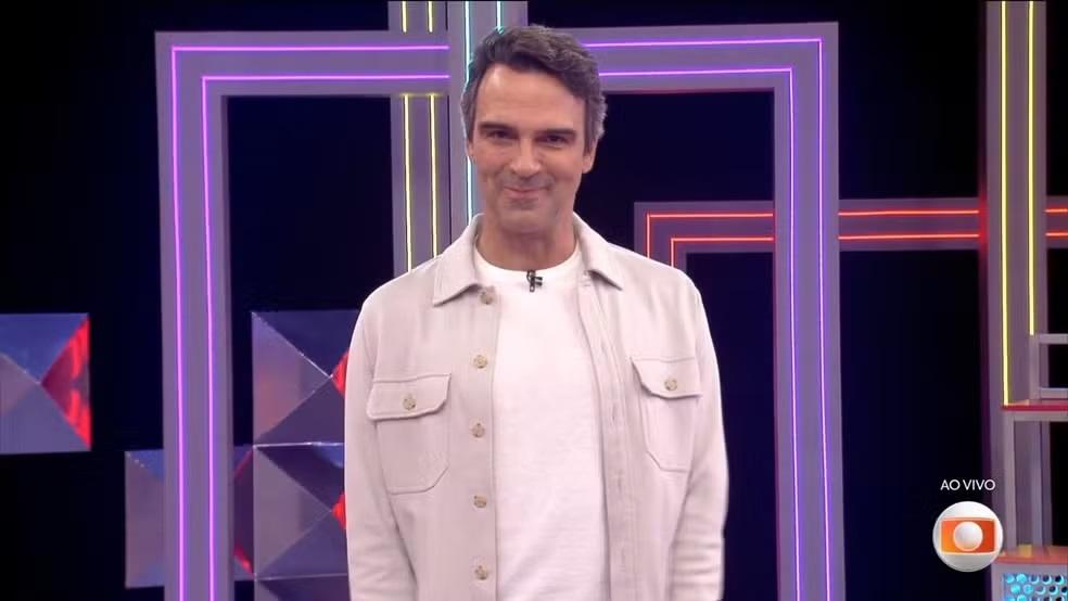 Tadeu Schmidt, apresentador do Big Brother Brasil, sorrindo e usando uma camisa clara sobre uma camiseta branca, em um cenário de estúdio com luzes geométricas roxas e brancas.
