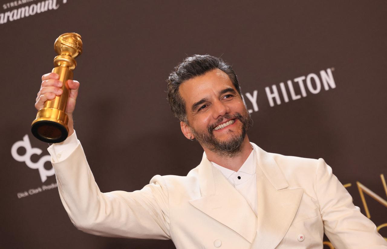 O ator Wagner Moura sorri radiantemente enquanto segura o troféu do Globo de Ouro com a mão direita elevada. Ele veste um elegante terno branco sobre uma camisa social clara, posando contra um fundo marrom com logotipos do evento em uma composição que celebra sua conquista.