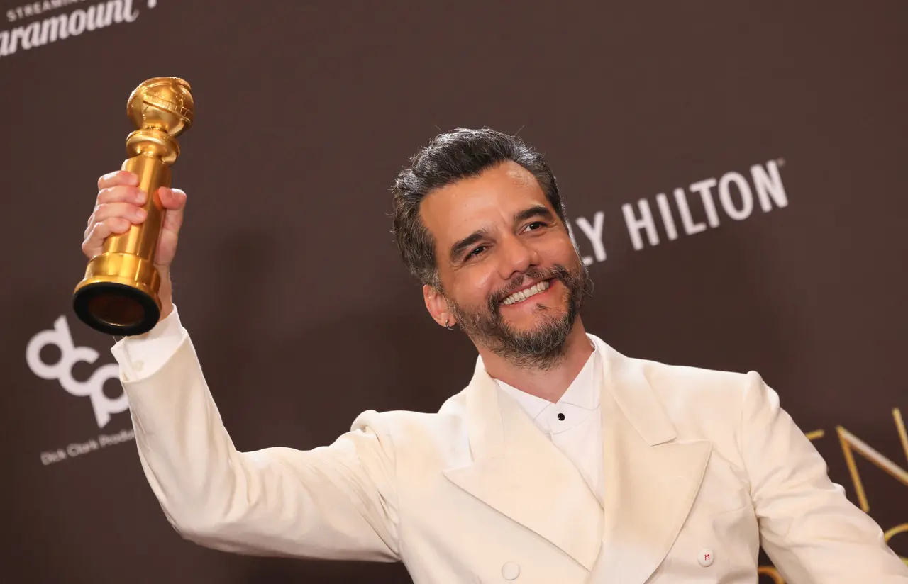 O ator Wagner Moura sorri radiantemente enquanto segura o troféu do Globo de Ouro com a mão direita elevada. Ele veste um elegante terno branco sobre uma camisa social clara, posando contra um fundo marrom com logotipos do evento em uma composição que celebra sua conquista.