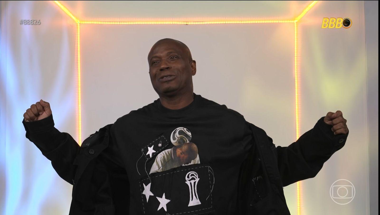 Edílson Capetinha celebra no programa BBB, usando uma camisa preta com estampa de uma figura segurando um troféu, em um palco iluminado por luzes neon.