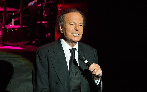 O cantor Julio Iglesias no palco, segurando um microfone e usando um terno preto.