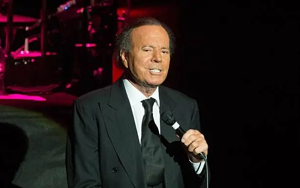 O cantor Julio Iglesias no palco, segurando um microfone e usando um terno preto.