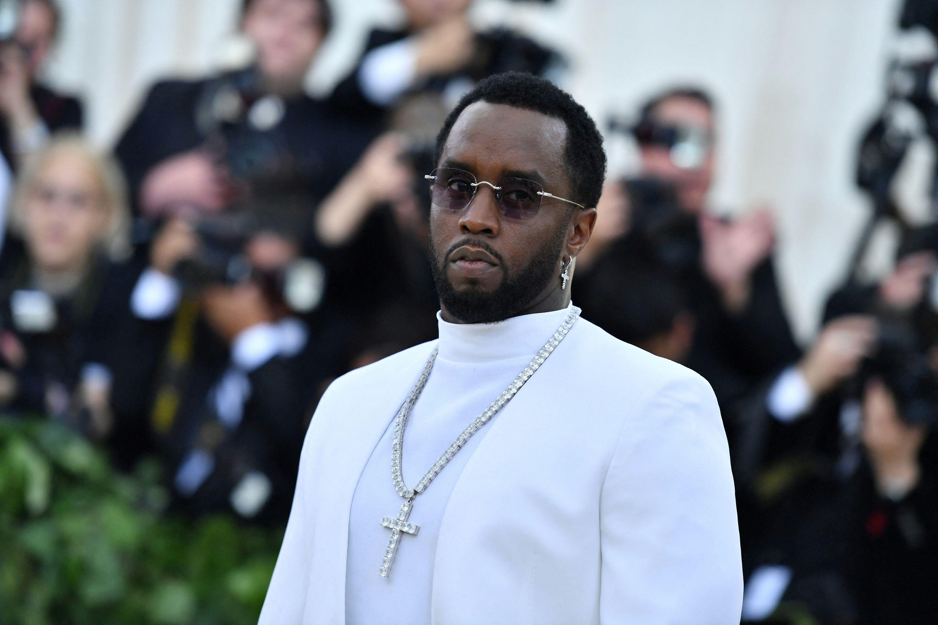 Sean Diddy Combs em um terno branco elegante, óculos escuros e colar de cruz, posando em um evento formal.