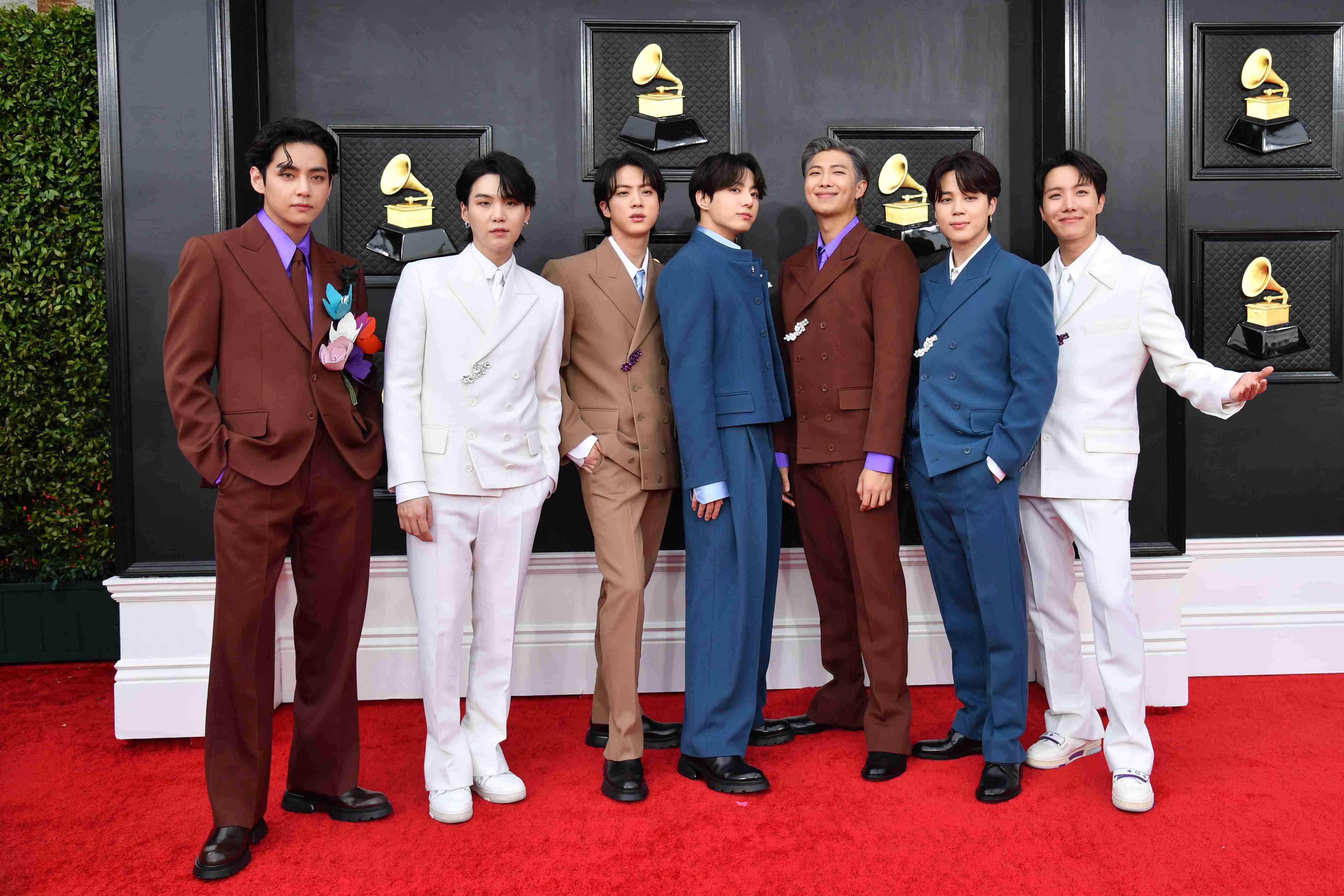 Sete membros do grupo K-pop BTS posam no tapete vermelho do Grammy, vestidos com ternos de cores variadas como marrom, azul, branco e bege, com estatuetas de gramofone douradas ao fundo.