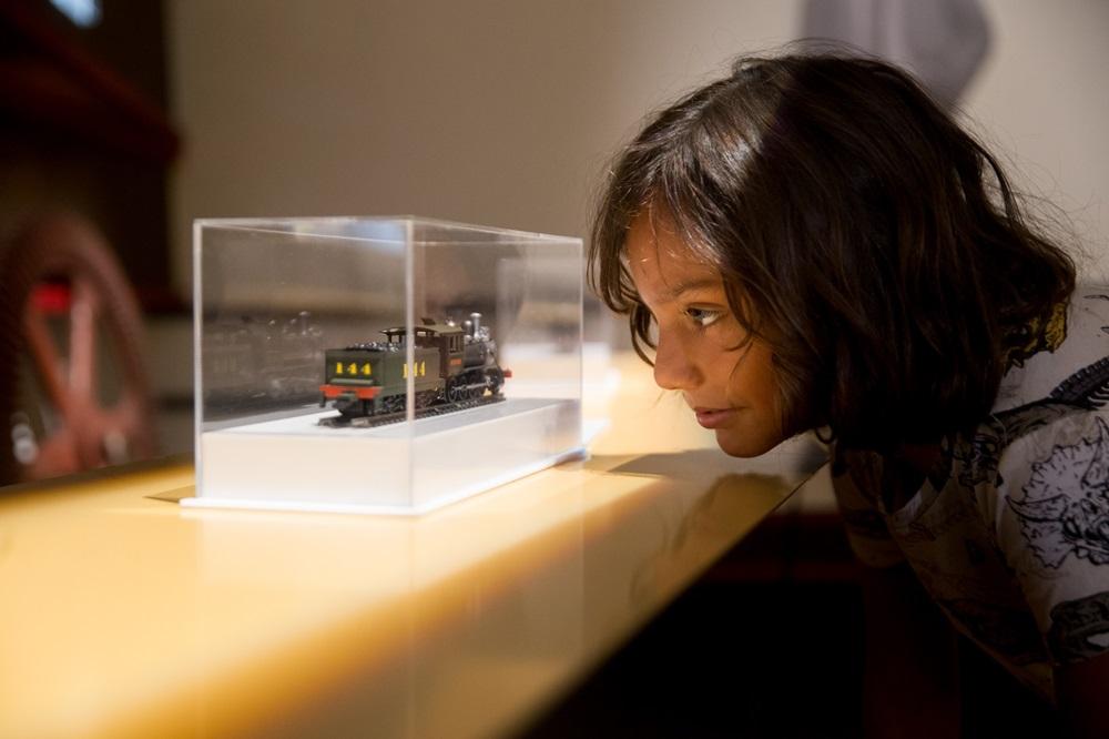 Na imagem, em close-up lateral, uma criança com expressão curiosa e concentrada olhando para um modelo reduzido de trem. A criança tem pele clara e cabelos castanhos ondulados na altura dos ombros. A miniatura do trem, de cor verde escura com o número 