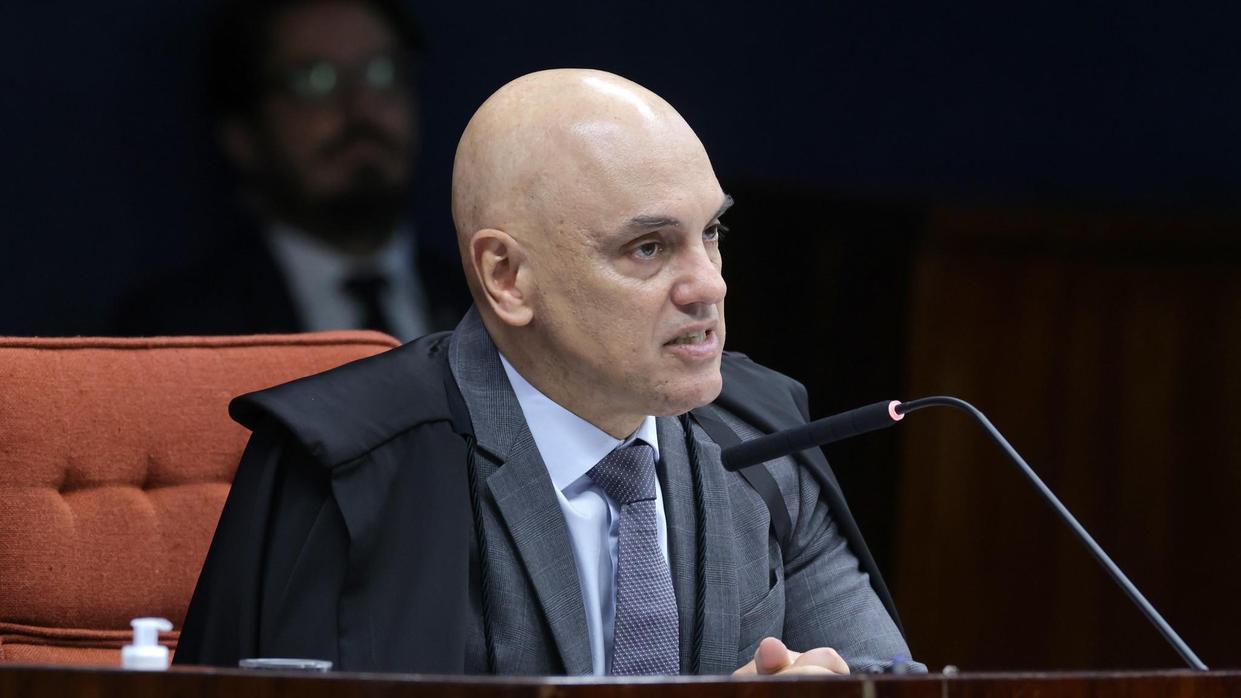 foto do ministro Alexandre de Moraes, sentado em bancada durante uma sessão formal, vestindo toga sobre terno e gravata, falando ao microfone em um ambiente institucional com mesa de madeira e cadeira estofada ao fundo, em contexto de julgamento ou audiência.