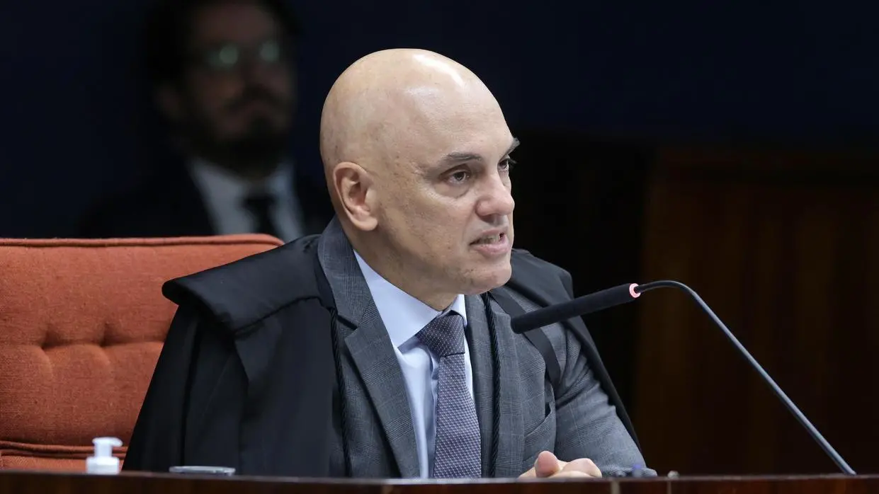 foto do ministro Alexandre de Moraes, sentado em bancada durante uma sessão formal, vestindo toga sobre terno e gravata, falando ao microfone em um ambiente institucional com mesa de madeira e cadeira estofada ao fundo, em contexto de julgamento ou audiência.