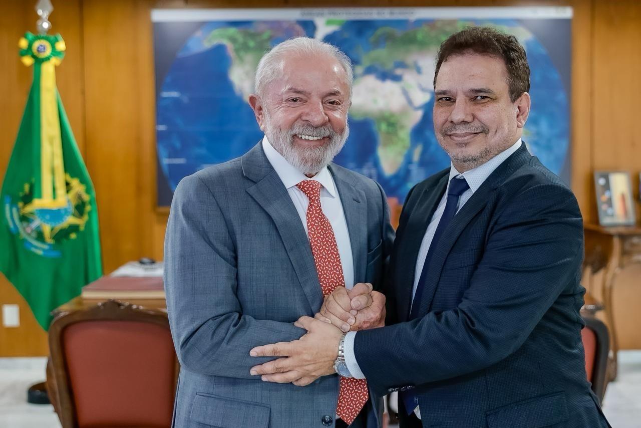 Presidente Luiz Inácio Lula da Silva e o novo ministro da Justiça Wellington César Lima e Silva apertam as mãos e sorriem em um escritório, com a bandeira do Brasil e um mapa-múndi ao fundo.
