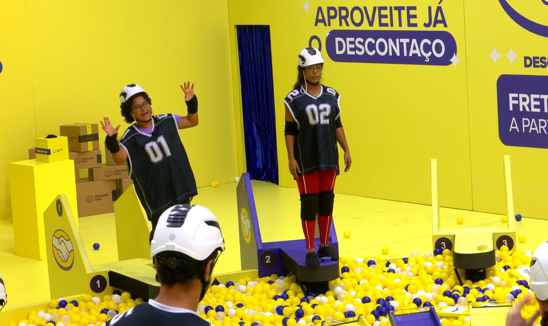 Sol e Milena, do BBB 26, com capacetes e camisetas esportivas em uma sala amarela vibrante cheia de bolas amarelas e brancas, em um cenário de game show.