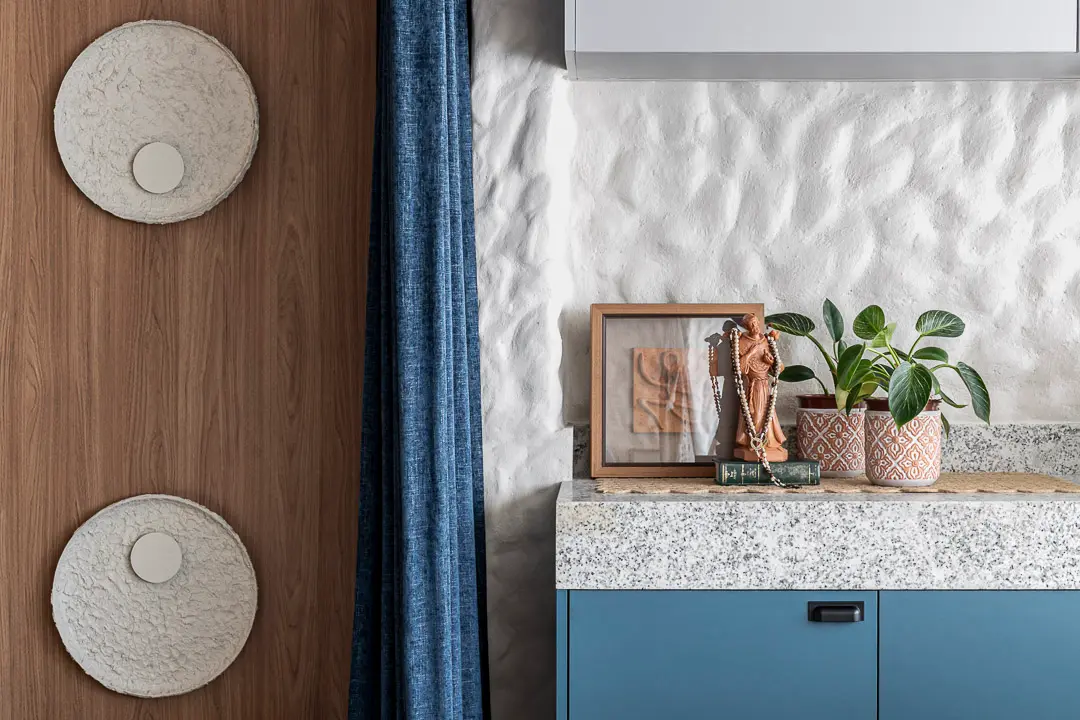 Uma parede branca com textura rústica e ondulada compõe o cenário ao lado de um armário azul com bancada de granito, onde repousam plantas e objetos decorativos. Painéis de madeira com arandelas circulares e uma cortina azul completam o ambiente, criando uma composição harmoniosa de texturas e cores serenas.