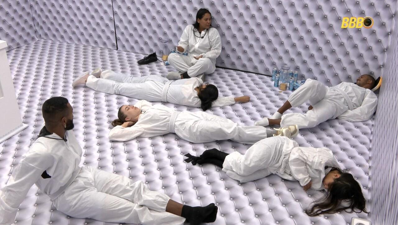 Seis pessoas de branco deitadas e sentadas em quarto branco, trajando roupas brancas.