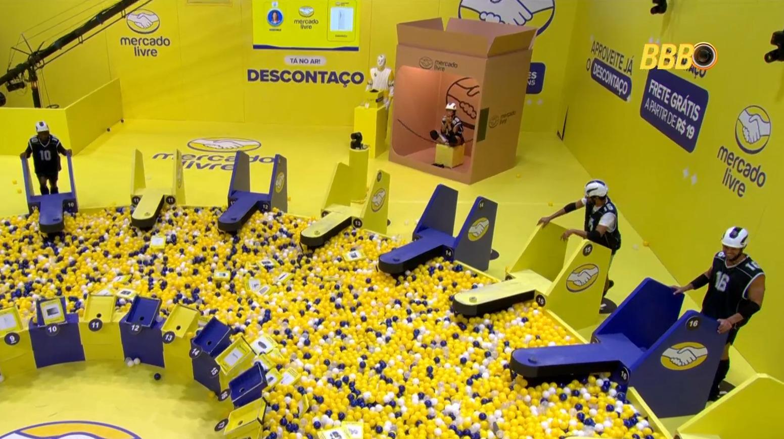 Vista aérea de quem ainda estar na competição da prova do líder no BBB 26, onde os participantes buscam itens em uma vasta piscina de bolinhas amarelas e azuis. O cenário, dominado pela cor amarela, exibe a marca Mercado Livre e estruturas que simulam caixas e veículos de transporte, destacando a parceria da prova.