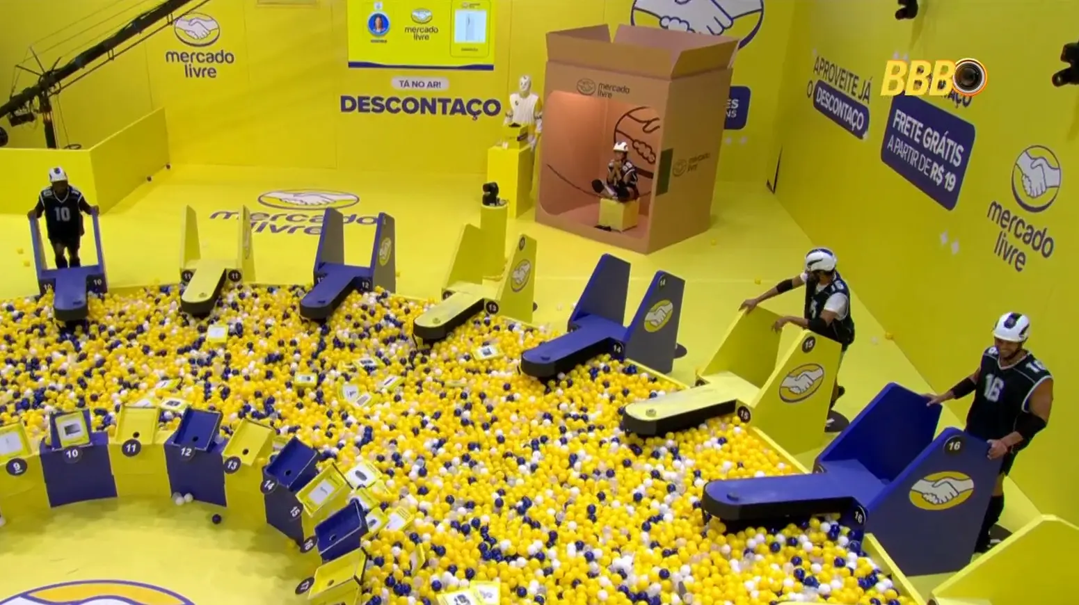Vista aérea de quem ainda estar na competição da prova do líder no BBB 26, onde os participantes buscam itens em uma vasta piscina de bolinhas amarelas e azuis. O cenário, dominado pela cor amarela, exibe a marca Mercado Livre e estruturas que simulam caixas e veículos de transporte, destacando a parceria da prova.