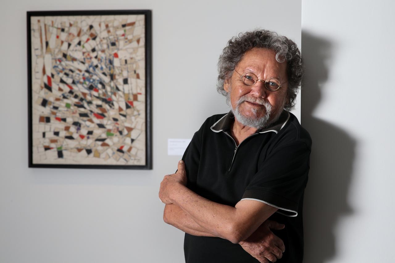 O artista visual Zé Tarcísio posa de braços cruzados em frente a uma obra do pintor Antonio Bandeira. Ele vestindo uma camisa polo preta e óculos redondos. O retrato foca em seu semblante sereno e cabelos grisalhos, com uma iluminação suave que destaca sua expressão contra a parede branca da galeria.