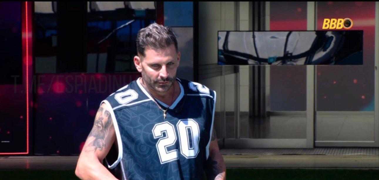 Homem com barba e camisa regata azul-marinho número 20, com tatuagens nos braços, em ambiente interno do Big Brother Brasil (BBB).