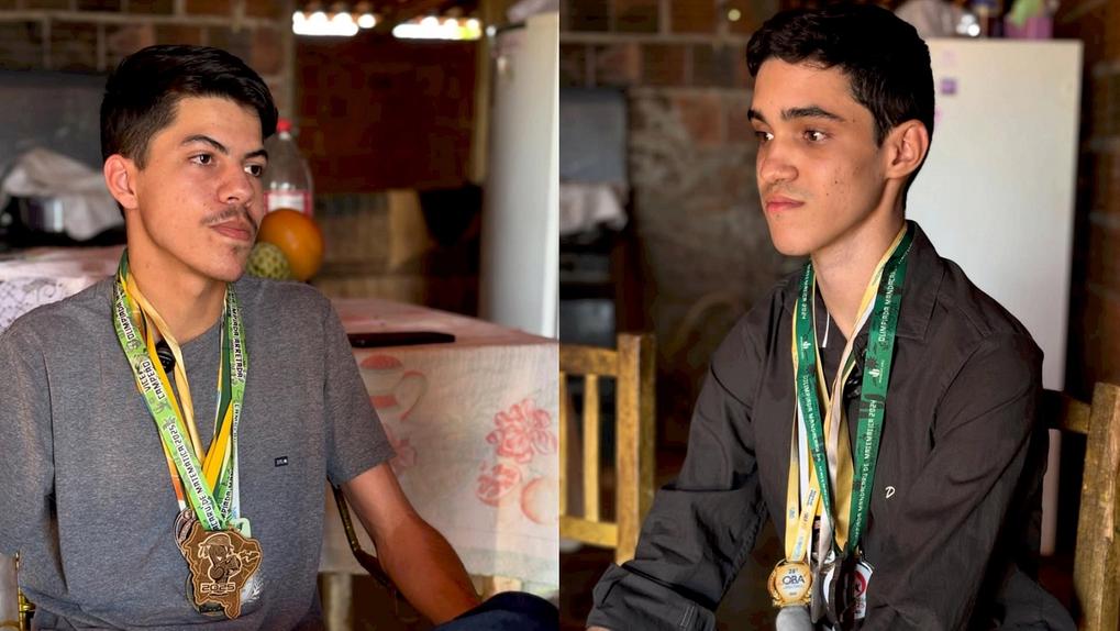 Imagem de dois rapazes jovens, Davi à esquerda e Willyan à direita, um usando uma camiseta cinza e o outro uma camisa escura, ambos exibindo medalhas penduradas no pescoço, em um cenário de casa do interior simples.