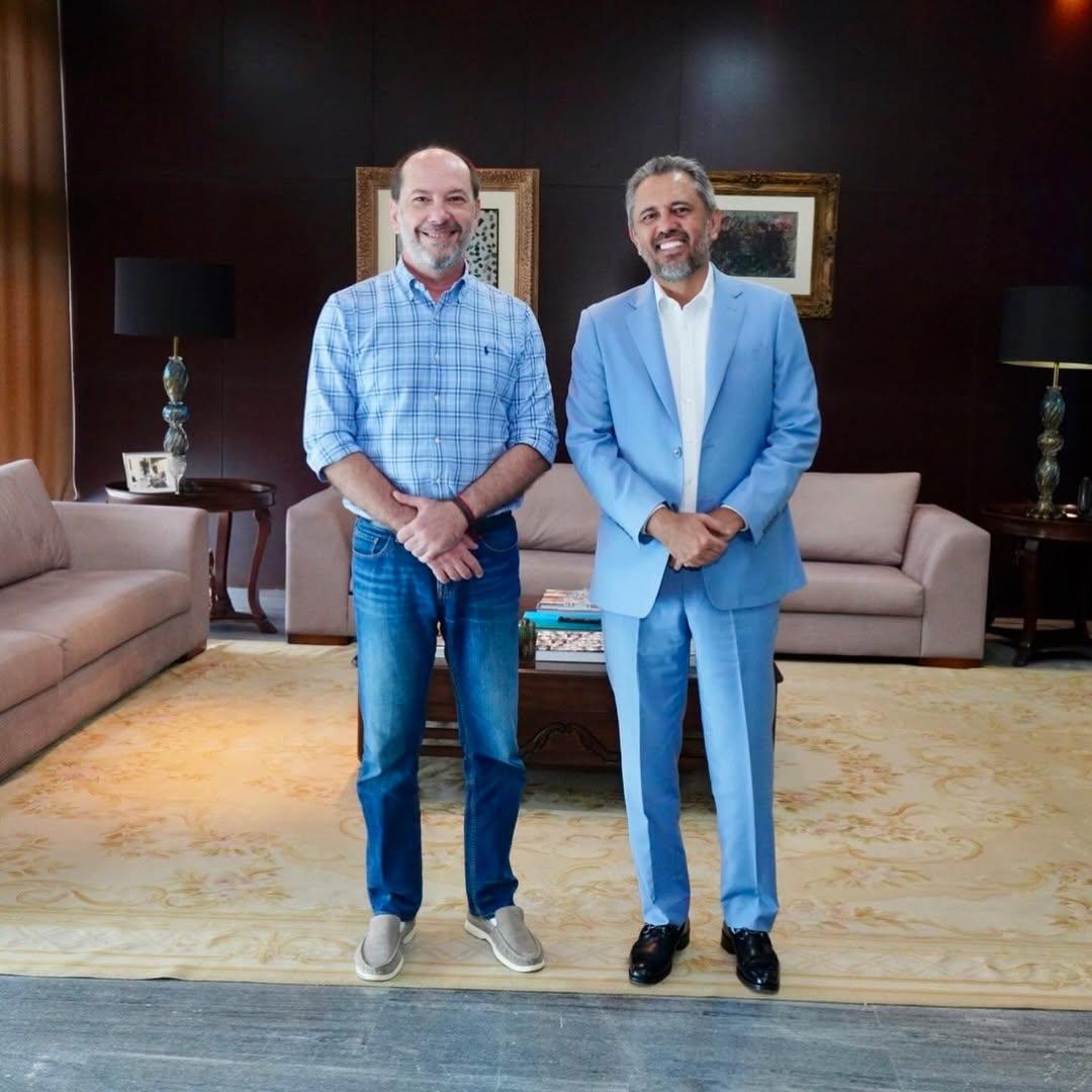 Ivo e Elmano, no gabinete do Governador. Ivo veste camisa xadrez azul e jeans; Elmano usa terno azul-claro com camisa branca. Ao fundo, sofás claros, mesas laterais com abajures e quadros na parede.