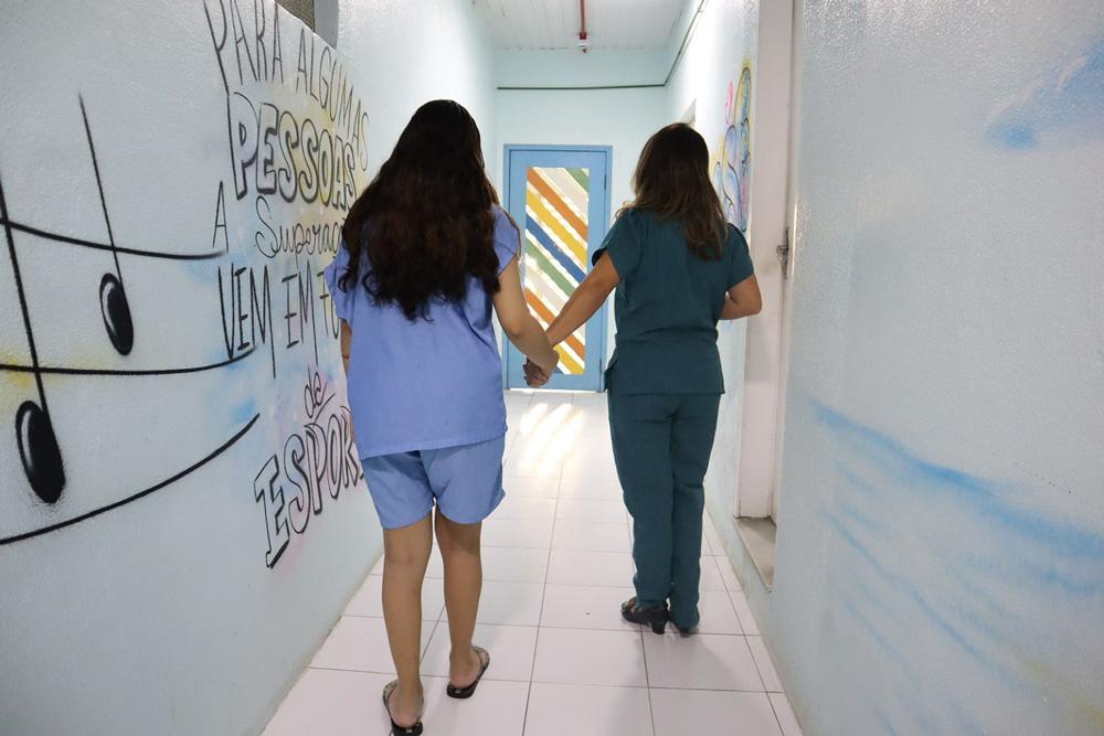 Profissional de saúde e paciente adolescente caminham de mãos dadas em corredor da ala de psiquiatria infantojuvenil do hospital SOPAI. Murais coloridos decoram as paredes laterais.