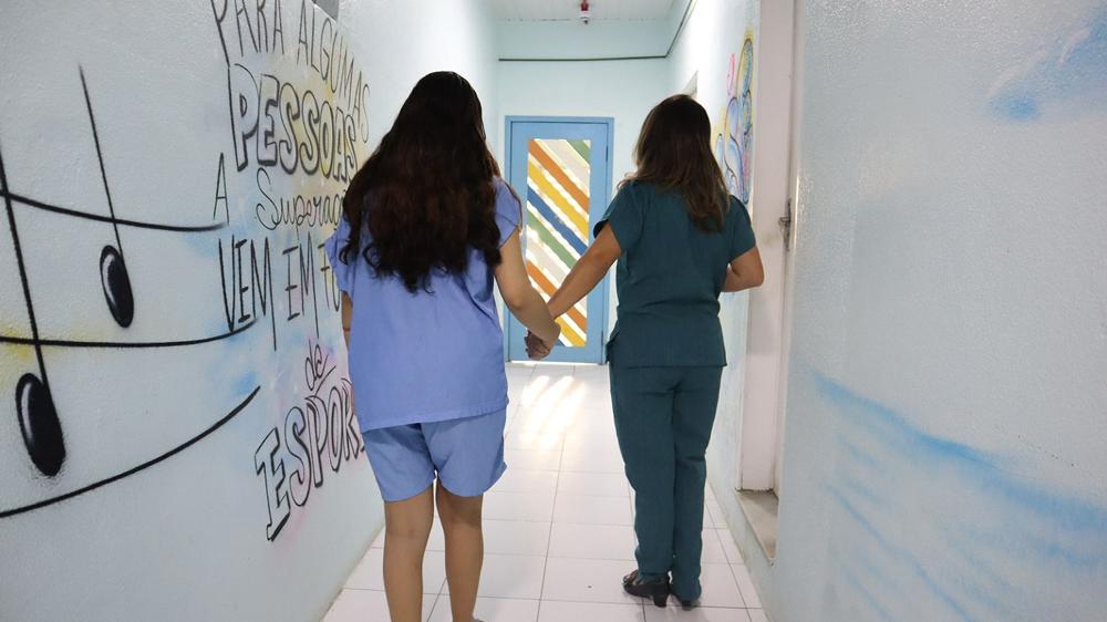 Profissional de saúde e paciente adolescente caminham de mãos dadas em corredor da ala de psiquiatria infantojuvenil do hospital SOPAI. Murais coloridos decoram as paredes laterais.