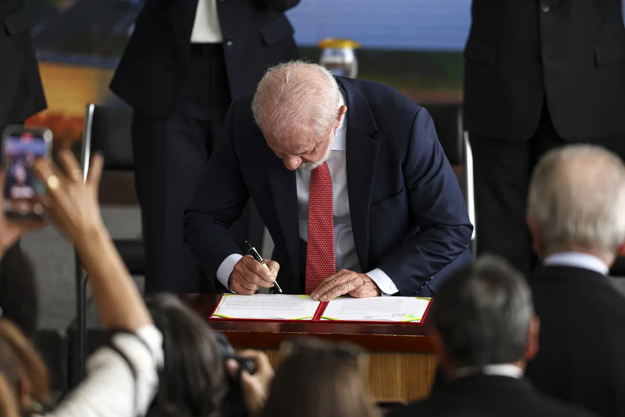 Imagem Lula, um homem idoso, assinando um documento oficial, com várias pessoas ao redor registrando o momento. Ele usa um terno azul marinho com roupa branca e gravata vermelha.