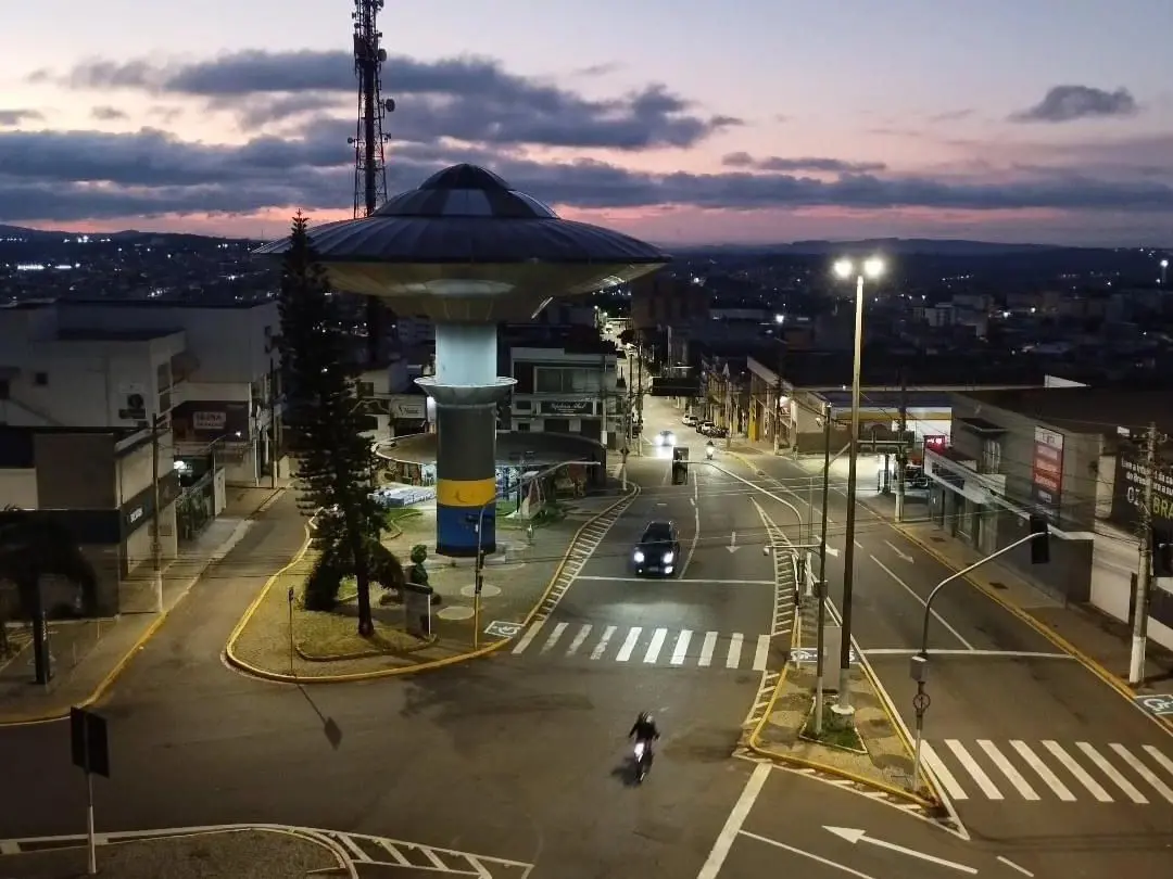 Imagem aérea da Nave do ET, uma caixa d'água em formato de disco voador no centro da cidade de Varginha, em Minas Gerais. Ele fica circulado por ruas com faixas de pedestres e residências. Ao fundo, há um céu em tons de laranja e roxo ao entardecer.