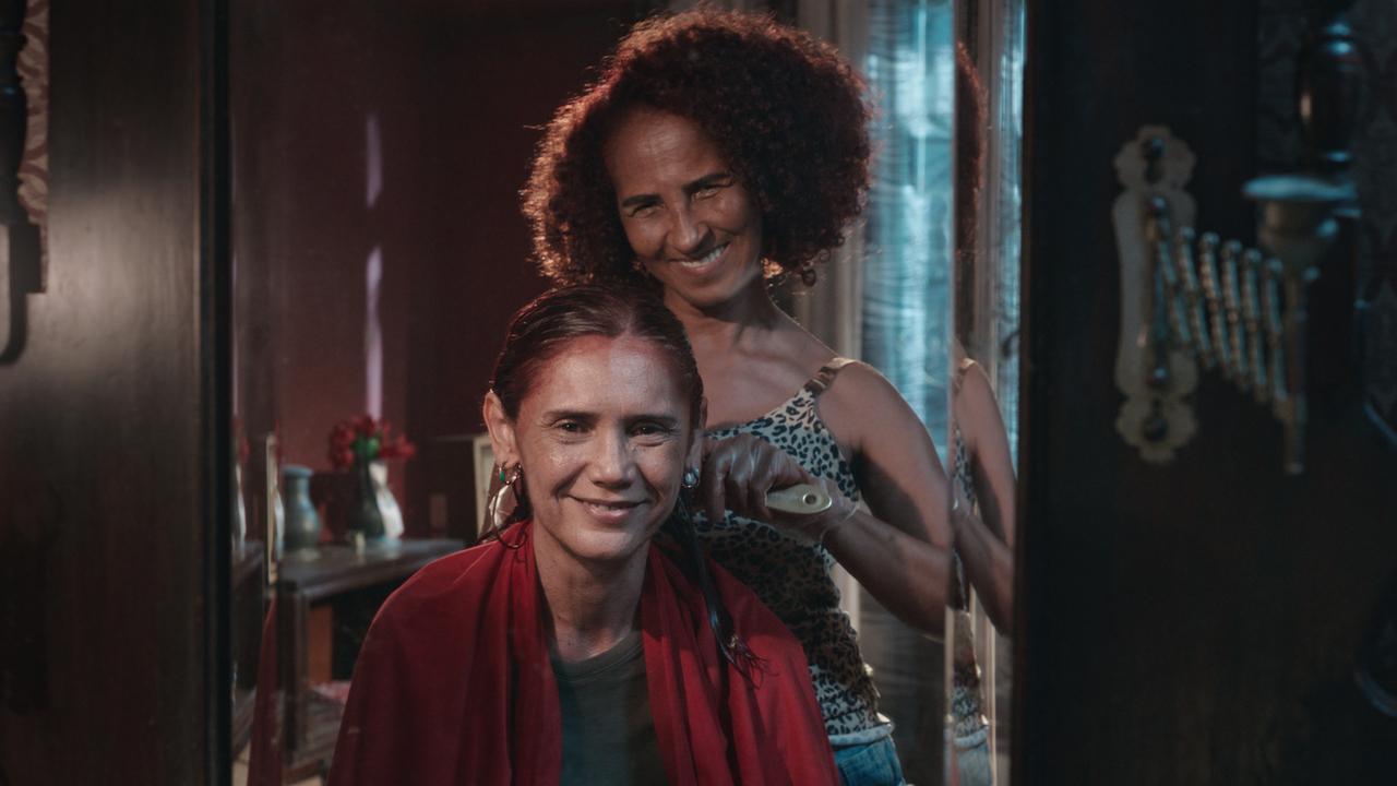 As atrizes Verônica Cavalcanti e Luciana Souza sorriem enquanto uma penteia o cabelo da outra em frente a um espelho de moldura escura. A cena possui uma iluminação suave e tons quentes, destacando a cumplicidade entre as personagens em um ambiente acolhedor e íntimo.