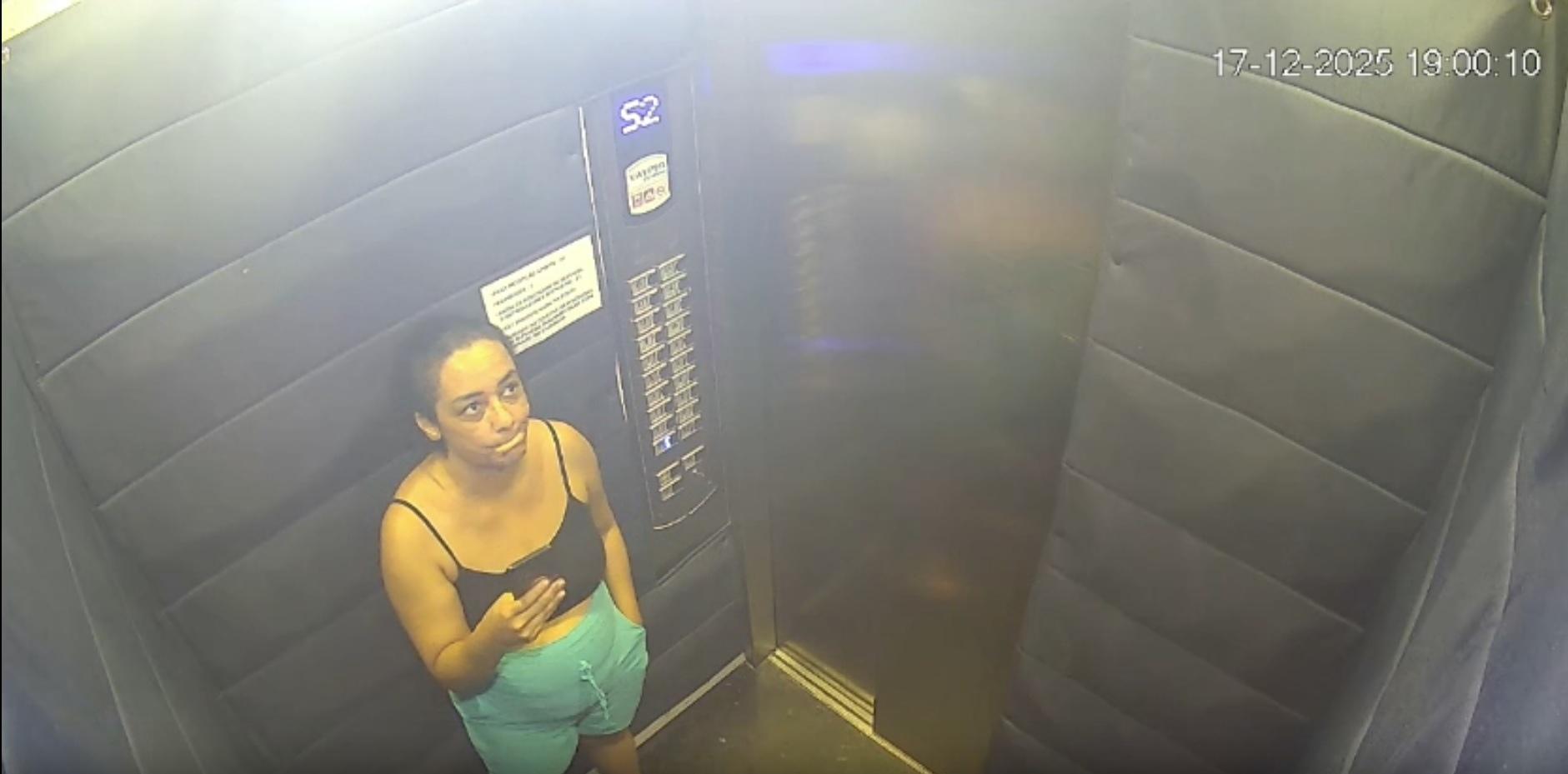 Imagem de câmera de segurança mostrando uma pessoa em um elevador acolchoado, vestindo blusa preta sem mangas e shorts azul‑claro, olhando para um telefone que segura com uma das mãos. A pessoa está encostada na parede lateral do elevador.
