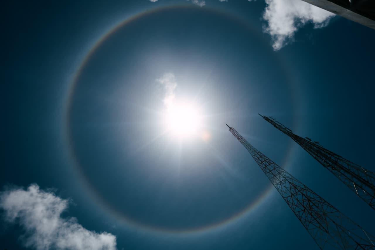 Círculo colorido similar a arco-íris ao redor do sol, com nuvens ao redor.