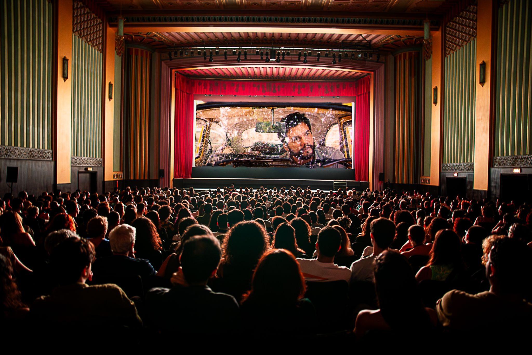 Cinema lotado, com cortinas vermelhas no fundo e exibição do filme O Agente Secreto, em Fortaleza.