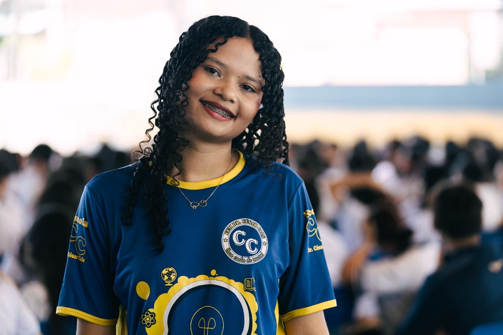 Retrato de Ana Rafaely sorrindo, com cabelos cacheados e usando aparelho. Ela veste uniforme escolar azul e amarelo. Ao fundo, outros estudantes aparecem desfocados em um ambiente amplo.