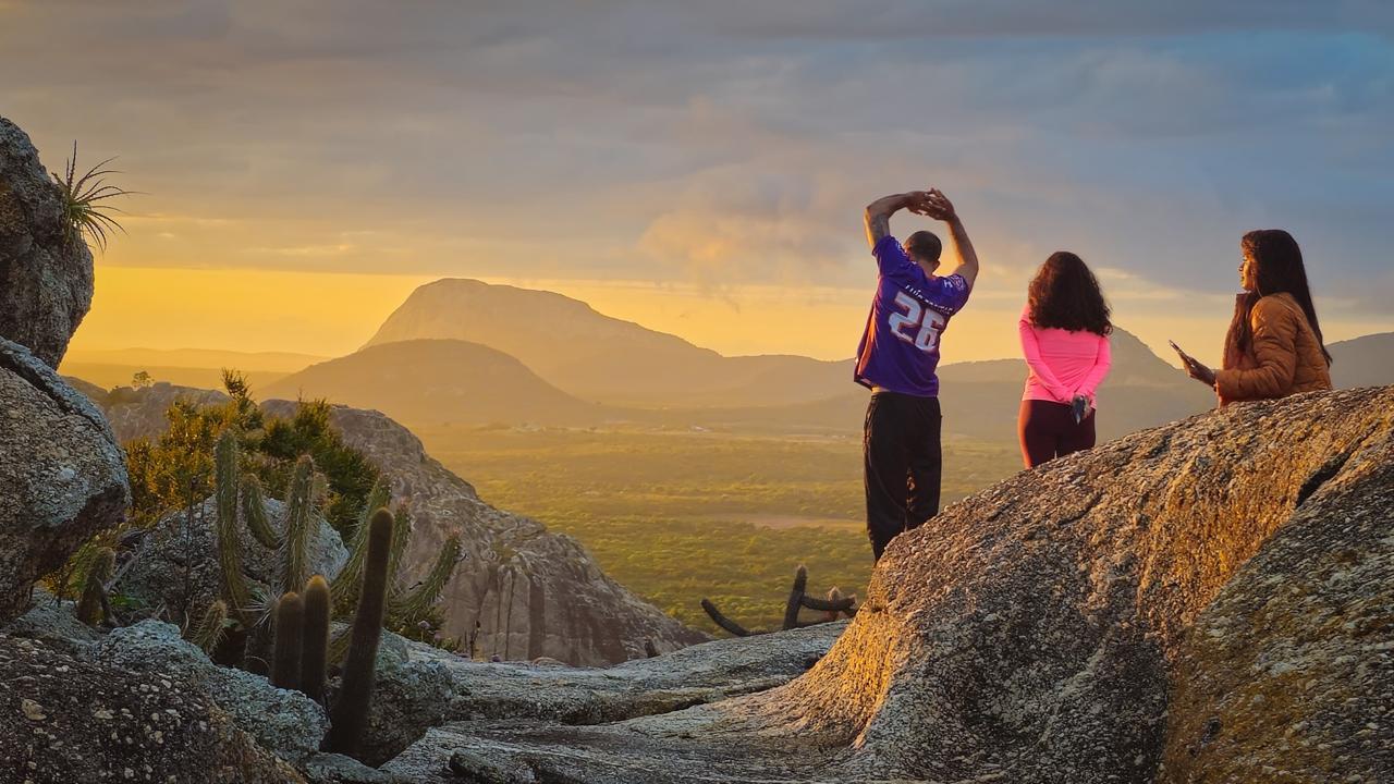Três pessoas admirando o nascer do sol em Quixadá, no Ceará, diante de um cenário de natureza, com montanhas ao fundo, rochas e cactos ao redor, capturadas ao entardecer.