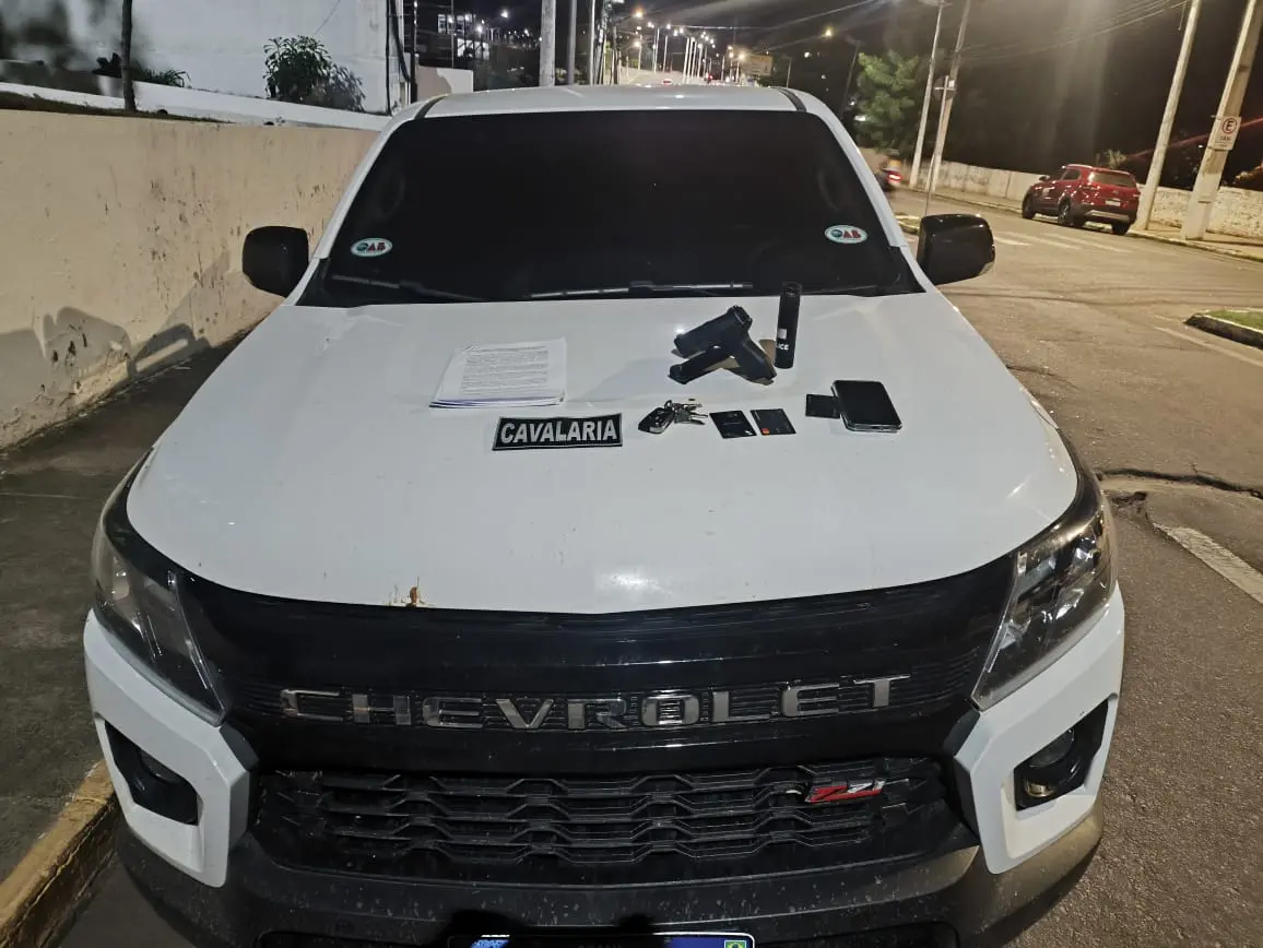 A imagem mostra um carro branco, com adesivos da OAB-CE, e em cima do capô há cartões bancários, uma chave de carro, um celular, um simulacro de arma de fogo e um spray de pimenta.