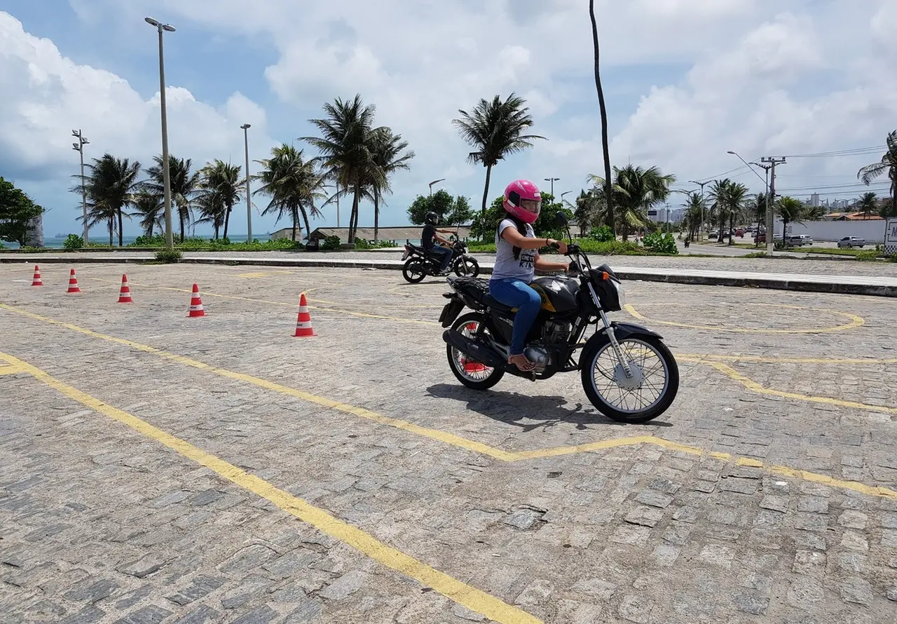Uma mulher de capacete rosa e jeans pilota uma motocicleta preta em um curso de treinamento ao ar livre, com cones laranja, linhas amarelas no chão de paralelepípedos, e coqueiros com o mar ao fundo na cidade de Fortaleza.
