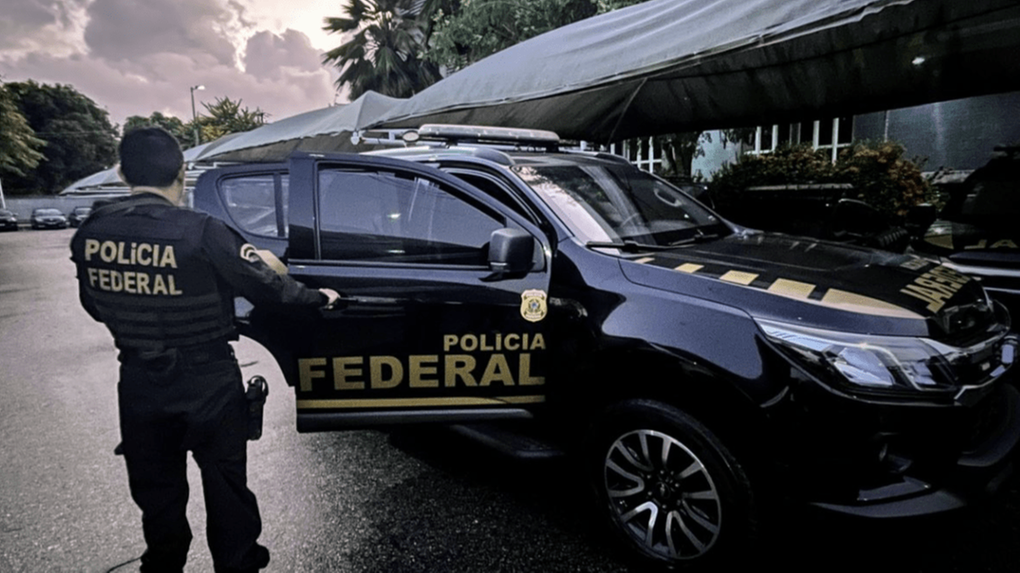 A imagem mostra um policial federal, fardado e de costas, ao lado de uma viatura da Polícia Federal, preta e adesivada.