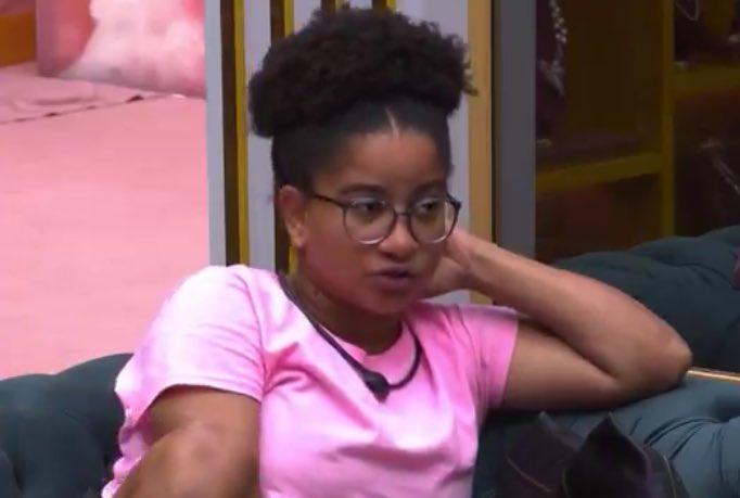 Milena, do BBB 26, com camisa rosa, sentada em sofá azul petróleo, próxima de espelho disposto em uma parede rosa.